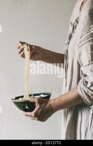 Frau in weißem Kleid Holding in den Händen Schüssel der Asiatischen Reis und Nudeln Nudelsuppe mit Stäbchen. Graue Wand als Hintergrund. Abendessen im asiatischen Stil. Stockfoto