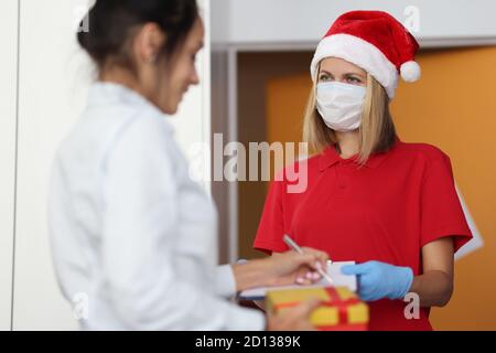 Kurierin Frau in roten weihnachtsmann Hut und schützende medizinische Maske auf Gesicht gibt dem Kunden zu unterzeichnen Dokument und Geschenk Stockfoto