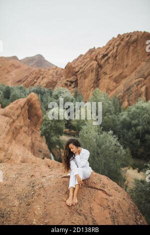 Hübsche Brünette Frau in weiß sitzen und genießen Blick auf Todra Schlucht Canyon in Marokko. Harmonie mit der Natur und Freiheitskonzept. Reiselebensstil. Stockfoto