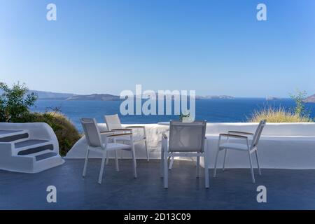 Vier weiße Stühle mit weißem Tisch auf der weißen Terrasse mit Blick auf das blaue Meer und die Caldera an einem sonnigen Tag, ohne Menschen. Oia, Santorini Insel, Griechenland Stockfoto