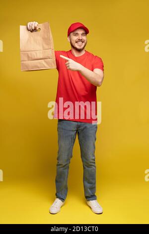 Mann in roter Mütze, T-Shirt mit Fast-Food-Bestellung isoliert auf gelbem Hintergrund. Männlicher Mitarbeiter Kurier halten leere Papierpaket mit Essen Stockfoto