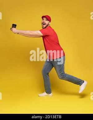 Lieferung Mann in roter Kappe blank T-Shirt Uniform isoliert auf gelbem Hintergrund. Ein Angestellter hält eine Tasse Kaffee zum Mitnehmen bereit. Stockfoto