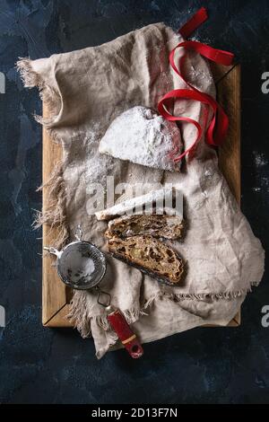 In Scheiben geschnitten Christmas Cake, traditionelle deutsche Festliches backen. Vollkorn Stollen mit Rosinen und Zucker Pulver auf Leinen Servietten, Holz- fach, Sieb, rot Ri Stockfoto