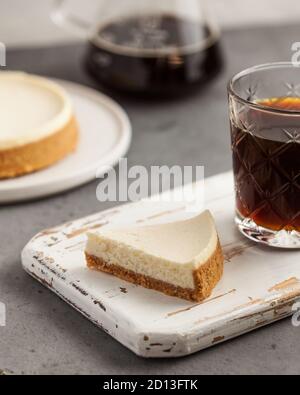 Nahaufnahme einer dreieckigen Scheibe Käsekuchen auf einem weißen Holztablett. Eine Kaffeekanne, eine Tasse Kaffee und Käsekuchen. Stockfoto