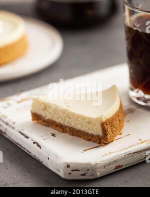 Nahaufnahme einer dreieckigen Scheibe Käsekuchen auf einem weißen Holztablett. Eine Kaffeekanne, eine Tasse Kaffee und Käsekuchen. Stockfoto