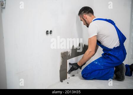 Der Generalarbeiter befestigt die Fliesen mit Zement an der Wand. Er trägt blaue Arbeitskleidung und schwarze Handschuhe Stockfoto