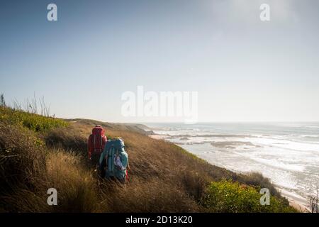 Zwei Wanderer gehen den Weg entlang der Dünen und Klippen mit hoher Vegetation, stehen neben einer Strandlandschaft am späten Abend Stockfoto