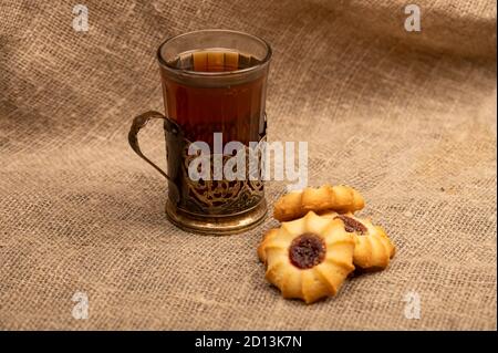 Hausgemachte Gebäck-Kekse mit Marmelade und einem facettierten Glas Tee in einem Vintage-Cup-Halter auf einem Hintergrund von homespun Stoff mit einer rauen Textur, close-up, Stockfoto