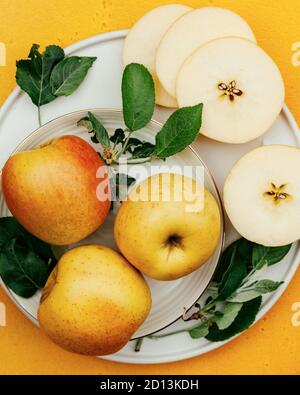 Rot-gelbe Äpfel mit grünen Blättern in ihrer Gesamtheit und geschnitten sind auf weißen Platten auf gelbem Hintergrund ausgelegt. Stockfoto