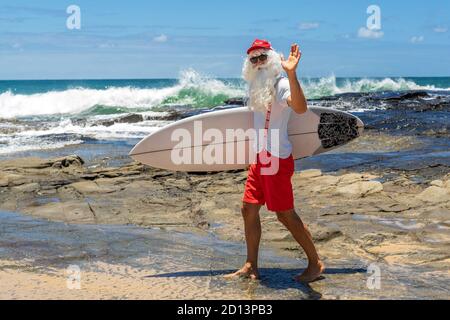 Weihnachtsmann mit einem Surfbrett mit dem Meer auf der Rückseite. Australien, Weihnachten im Sommer. Stockfoto