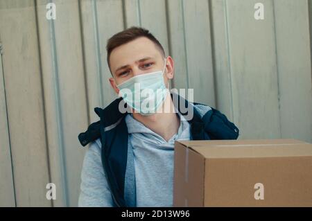 Junge Lieferung Kerl in covid Maske schaut auf die Kamera Und fasst eine Schachtel Stockfoto