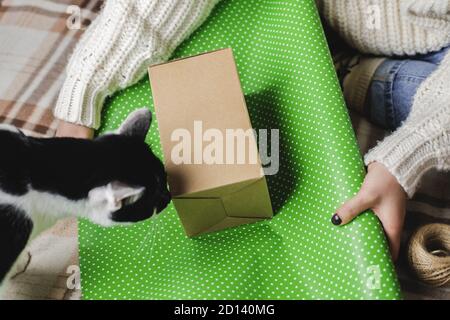 Junge Frau sitzt auf karierten in kuschelig gestrickten weißen Wollpullover, Socken und wickelt Weihnachtsgeschenk in Polka Punkt Geschenkpapier. Lustige schwarz-weiße Tuxe Stockfoto