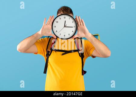 Time-to-Travel-Konzept. Junger Mann mit Campingausrüstung, die Uhr vor seinem Gesicht über blauem Studiohintergrund hält Stockfoto