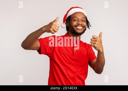 Lächelnder afro-amerikanischer Kerl mit Bart und Dreadlocks im weihnachtsmann Hut zeigt Daumen nach oben Blick auf die Kamera, mag Winterferien. Innenaufnahme im Studio Stockfoto