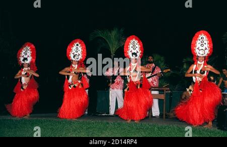 Vier lächelnde weibliche Entertainer treten in leuchtend roten Kostümen während eines Hawaii Luau auf, das einheimische Speisen und Tänze in einem Ferienresort auf der Insel Maui in Hawaii, USA, bietet. Ein Luau ist der beste Ort für Besucher, um traditionelle Hula-Tänze zu sehen, wo alte hawaiianische Geschichten mit den Händen und Armen der Tänzer erzählt werden. Tänze anderer polynesischer Kulturen sind oft Teil der Show, vor allem von Tahitian-otea-Tänzern, wie sie hier zu sehen sind. Sie sind leicht durch hohe aufwendige Kopfbedeckungen, Kokosnussschalen und große Grasröcke zu identifizieren, die mit den schnell schüttelnden Hüften der Mädchen vibrieren. Stockfoto