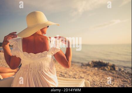 Pensionierte kaukasische Frau trägt Sonnenschutzkappe Entspannung auf einem Strand Sitz auf weißem Kunststoff-Liegestuhl. Urlaubszeitthema. Stockfoto