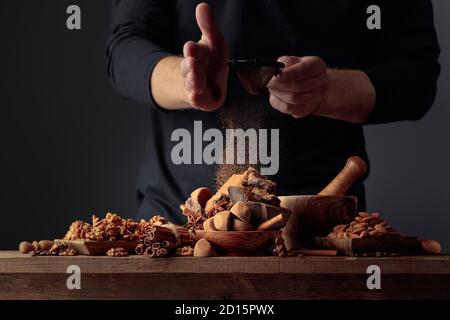 Der Koch streut die Schokolade mit Kakaopulver. Auf einem alten Holztisch Süßigkeiten, Kaffeebohnen, Zimt, Anis, Nüsse und Stücke von gebrochenem schwarzem Choco Stockfoto