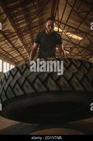 Harter männlicher Athlet, der eine Reifenflip-Übung macht. Muskulöser Mann, der im alten Lager Cross-Training macht. Stockfoto