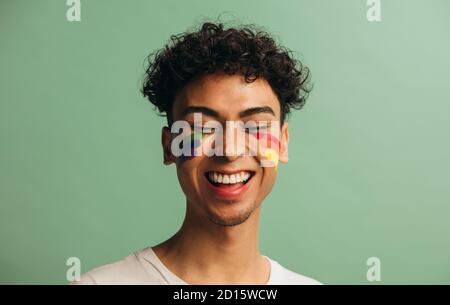 Nahaufnahme eines fröhlichen jungen Mannes mit einer stolzen Flagge auf dem Gesicht. Gay Mann mit Regenbogen Gesichtsfarbe lächelnd vor pastellgrünen Hintergrund. Stockfoto