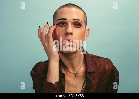 Geschlecht Flüssigkeit männlich mit Make-up Blick auf Kamera vor blauem Hintergrund. Gay Mann trägt Seidenhemd Anwendung Make-up blauen Hintergrund. Stockfoto