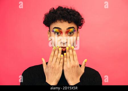 Gay Mann trägt Regenbogen farbigen Lidschatten und Smiley Gesicht auf Fingernagel auf rotem Hintergrund. Transgender männlich mit funky Make-up. Stockfoto