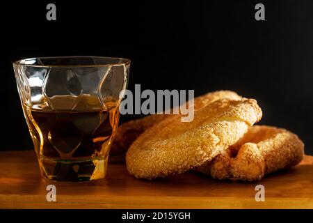 Nahaufnahme von süßen Donuts mit Zucker und einem Glas Rum auf Holzbrett und schwarzem Hintergrund. Stockfoto