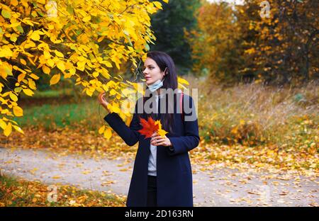 Frau mit abgesenkter Schutzmaske geht im Herbstpark. Zeit in der Natur während des Coronavirus-Ausbruchs. Stockfoto