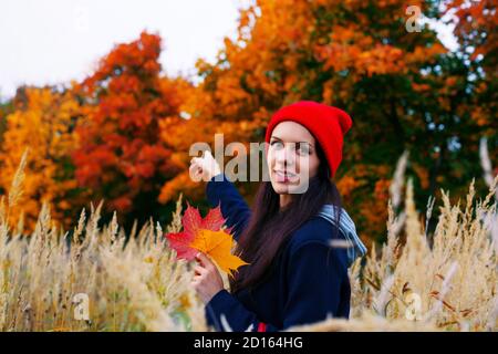 Lächelnde Frau in rotem Hut zeigt auf bunten Herbstwald hinter ihr. Herbstkonzept. Stockfoto