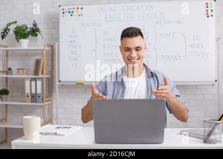 Englischlehrer zu Hause, Online-Praxis während COVID-19. Der lächelnde junge Mann freut sich über den Erfolg der Schüler in der Lektion im Inneren des Hauses mit der Tafel Stockfoto