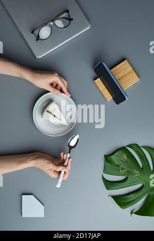 Minimal oben Ansicht flache Lage der Frau essen Käsekuchen über grauen Arbeitsplatz Hintergrund mit tropischen Blatt und Business-Accessoires, Kopierer Platz Stockfoto