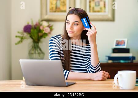 Attraktive Frau hält Kreditkarte in der Hand, während sie hinter ihrem Laptop sitzt und träumt. Stockfoto