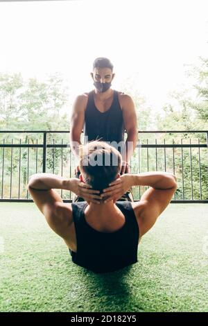 Trainer trägt Gesichtsmaske hilft jungen Mann Sit-ups zu tun Stockfoto