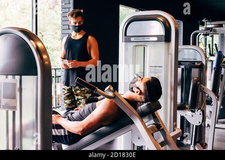 Trainer trägt eine Gesichtsmaske hilft einem jungen Mann tun Sein Training im Fitnessstudio Stockfoto