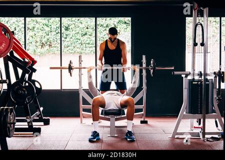 Personal Trainer trägt eine Gesichtsmaske hilft jungen Männern durchführen Ihre Übungen im Fitnessstudio Stockfoto