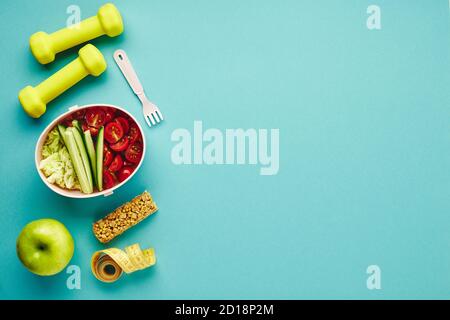 Kreative flache Lay von Sport- und Fitnessgeräten. Kurzhanteln, Maßband und Lunchbox mit gesundem Gemüsesalat auf hellblauem Hintergrund. Stockfoto