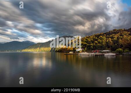 Herbstlicher Blick auf den Comer See, umgeben von Bergen, mit imposanten Sturmwolken nähern sich. Die Wälder mit warmen Herbstfarben spiegeln sich in der Stockfoto