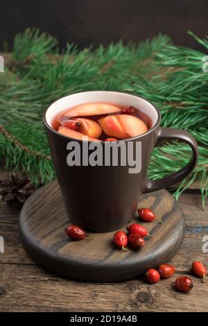 Weihnachtsgetränk in einer braunen Tasse mit Äpfeln, Beeren auf einem Hintergrund von Tannenzweigen auf einem dunklen Holzhintergrund. Stockfoto