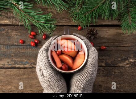 Weihnachtskomposition mit Händen in gestrickten Fäustlingen, die einen Becher mit einem heißen Getränk auf einem dunklen Holzhintergrund mit Beeren und Kiefernzweigen halten. Stockfoto
