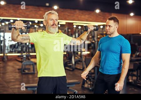 Training Körper in jedem Alter. Fokussierte reifen kaukasischen Mann in Sportbekleidung Training mit Hanteln unter Aufsicht eines jungen professionellen Personal Stockfoto