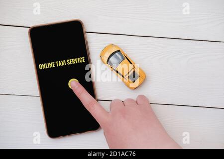 Weibliche Hand berühren Bildschirm-Taste, um Taxi rufen. Urban Online Taxi Service Anwendung. Gelbes Spielzeug Auto in der Nähe von Smartphone auf dem Tisch. Online-Kundenservice. Stockfoto