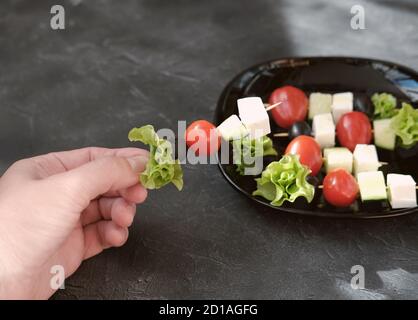 Rote Kirschtomaten, Mozzarella, Oliven und grüner Salat auf Spieße. Sommerappetitset. Rohe vegetarische Kebabs in der Hand der Frau Stockfoto