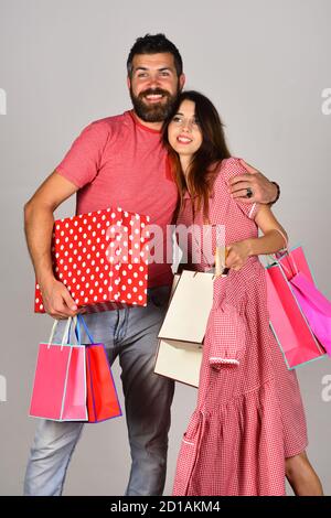 Kerl mit Bart und Mädchen mit lächelnden Gesichtern tun Einkaufen. Liebespaar hält Einkaufstaschen auf hellgrauem Hintergrund und Umarmungen. Einkaufs- und Ausgabenkonzept. Mann mit Bart hält Box. Stockfoto