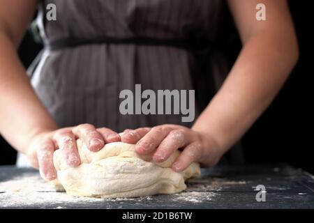 Frau Koch Kneten Teig für Brot oder Pizza. Bäckerei oder hausgemachtes Brot in Prozess, Frau in grau Schürze vor schwarzem Hintergrund. Konzept von Backen und Patisserie. Stockfoto