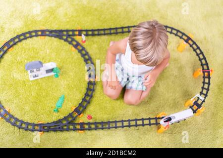 Kleiner Junge spielt mit Plastikeisenbahn. Kind mit Spielzeugzug. Pädagogisches Spielzeug für kleine Kinder. Kleiner Junge baut Eisenbahnschienen auf grünem Teppich. Stockfoto