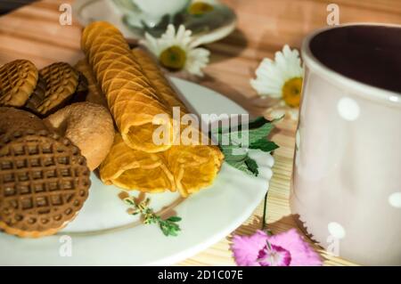 Waffelbrötchen und Kekse auf einem weißen Teller und eine Tasse Tee. Waffeln auf der Seite. Komposition auf einem hölzernen Hintergrund. Sichtbare Waffelstruktur Stockfoto