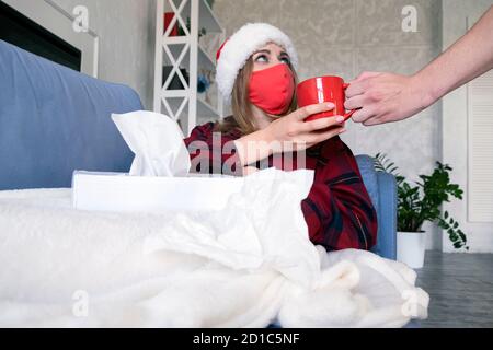 Gesundheit, Familie, Beziehung und Pflege Konzept. Mann gibt heißen Tee zu kranken jungen Frau auf dem Sofa in medizinischen Schutzmaske und weihnachtshut nach Hause liegen Stockfoto