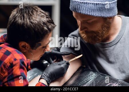 Ein Tattoo-Künstler mit kurzen Haaren und Piercings bereitet sich darauf vor, ein Tattoo auf den Arm eines Mannes in einem Tattoo-Salon zu bekommen. Stockfoto