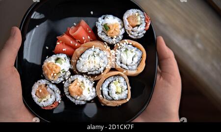 Hausgemachte Sushi-Rollen im asiatischen Stil. philadelphia und kalifornien Rollen auf einem schwarzen Teller mit mariniertem Ingwer. Menschliche Hände halten orientalische Vorspeise. Stockfoto
