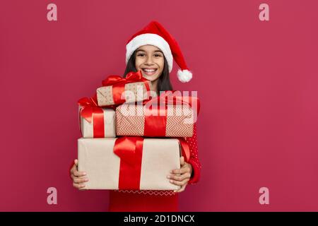 Happy indian Kind Mädchen tragen weihnachtsmann Hut hält viele Geschenke Boxen auf rotem Hintergrund. Stockfoto
