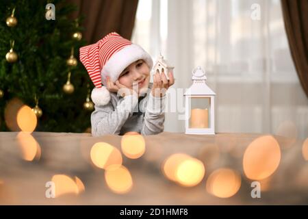 Eine Laterne, die auf ein dekoratives Haus schaut, ein lächelndes Kind, sitzt mit einem Spielzeug in der linken Hand und lehnte sich auf seine rechte Handfläche. Stockfoto